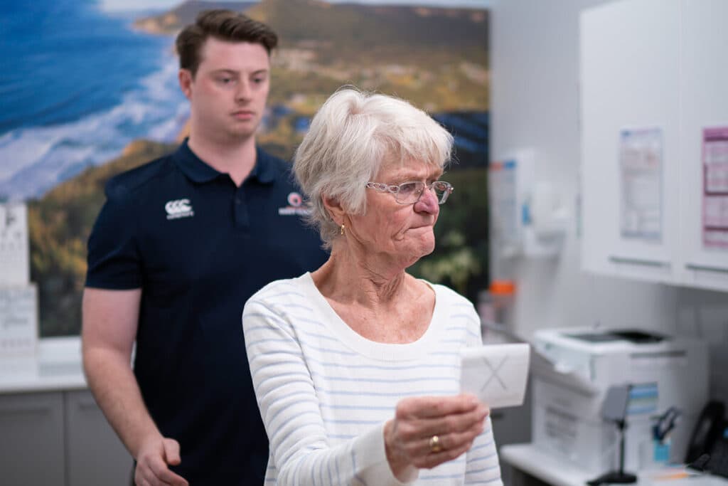 An older patient holding a vision target at arm's length during a balance exercise, with a Dizzy Clinics physiotherapist standing behind her